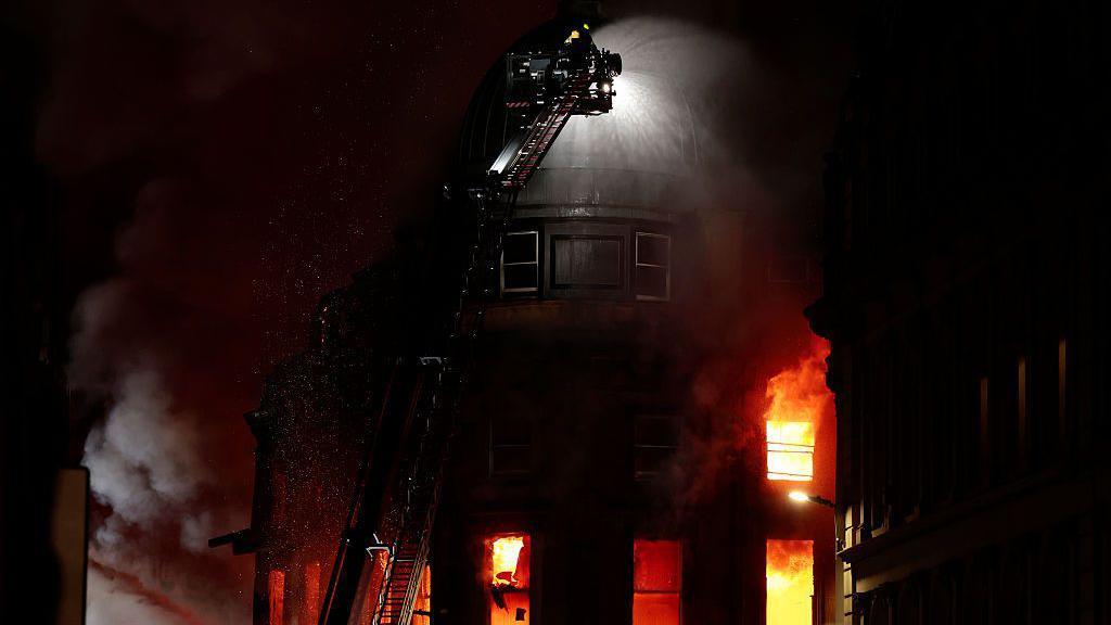 Një zjarr që shpërthen në një ndërtesë të lartë, me një zjarrfikës mbi një shkallë të zgjatshme që spërkat ujë sipër saj.