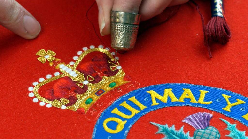 detail image of fingers near an intricately embroidered crown next to a circle with latin text and part of a thistle