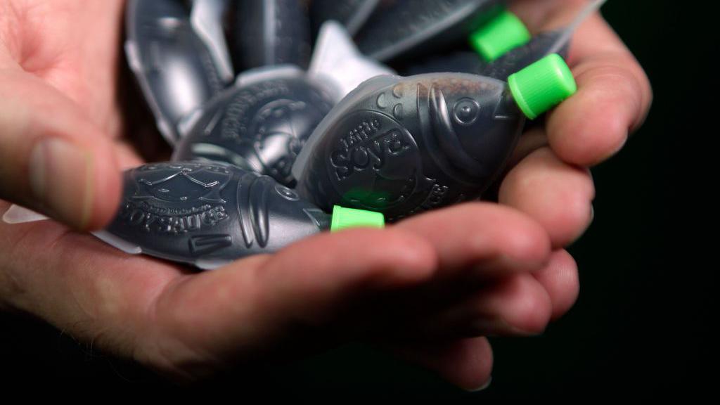 A close-view of hands holding several fish-shaped soy sauce dispensers with a green cap.