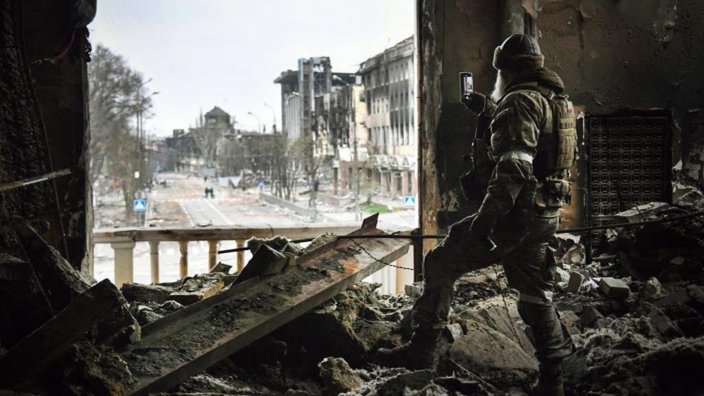 A Russian soldier stands in the ruins of Mariupol's theatre in spring 2022, taking a photo out of a destroyed window using his phone