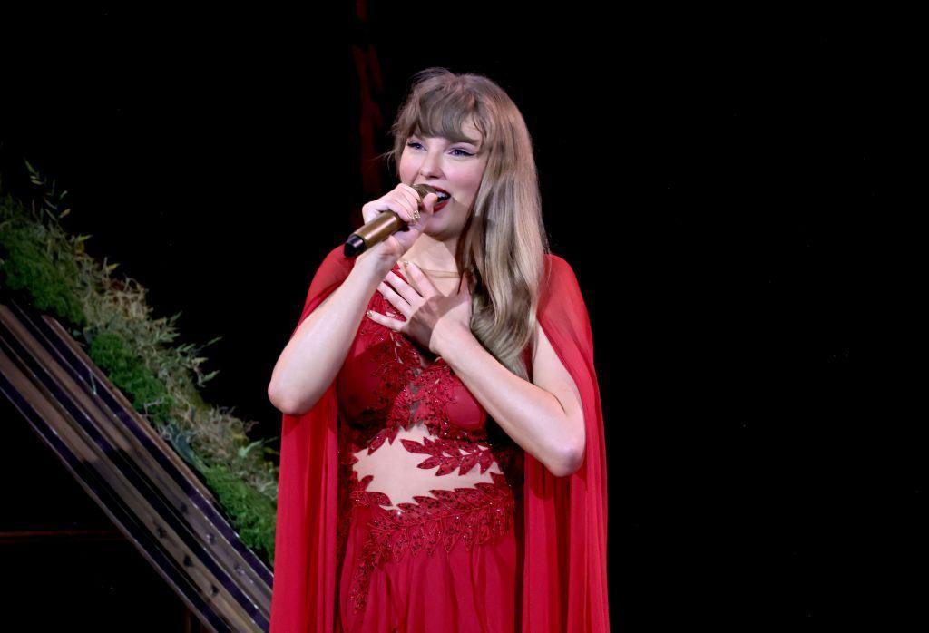 Taylor Swift onstage in a red dress and cape, during the Folklore/Evermore set of her Eras Tour