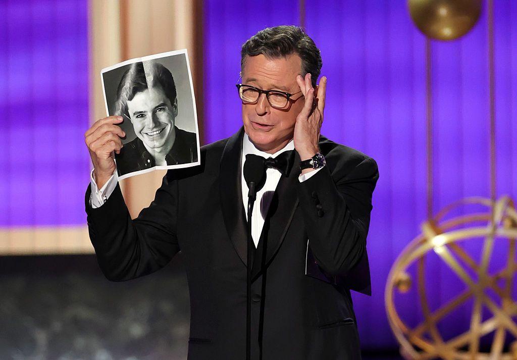 Stephen Colbert wearing a tuxedo puts his hand on his cheek and holds up his old headshot onstage at the Primetime Emmy Awards
