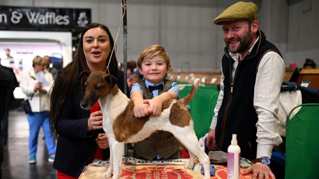 A woman with a black blazer and red dress stands next to a boy in a brown tweed suit and a man in a black waistcoat, white shirt and yellow hat. The boy is holding onto a brown and white fox terrier in front of him.