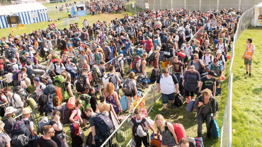 A huge crowd of people carrying luggage and tents into Glastonbury Festival. They are queuing up the grass between metal barriers, ready to go through security.