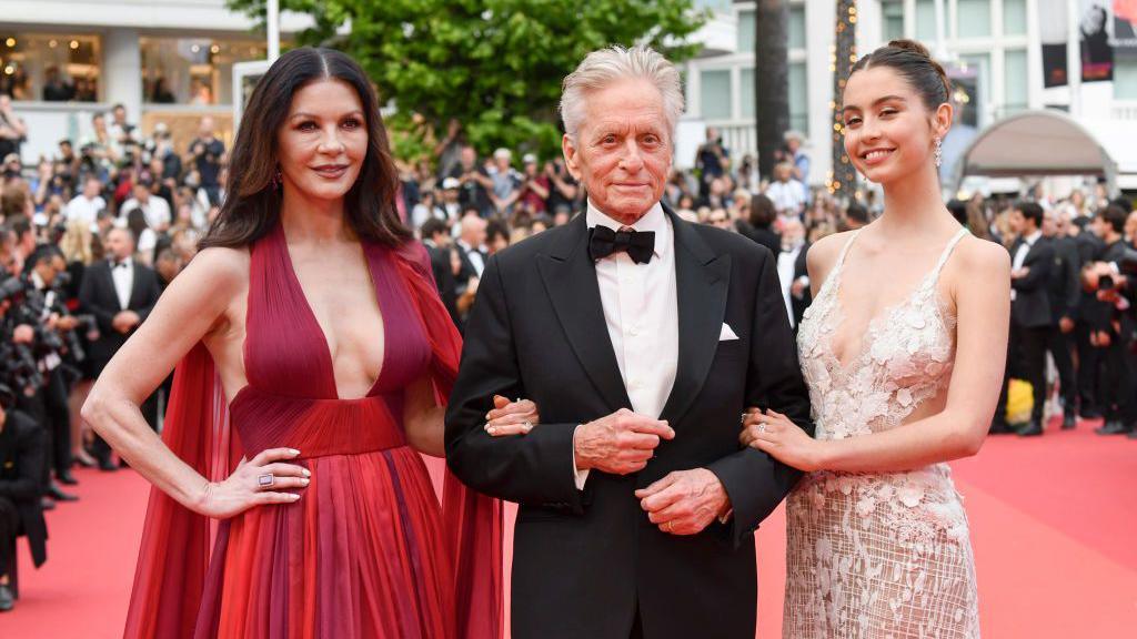 Carys Douglas, Michael Douglas and Catherine Zeta-Jones on the Cannes Film Festival Red Carpet. Michael is in the middle in a black tuxedo, with Carys and Catherine on either side, they are linking arms. Catherine Zeta Jones is wearing a long, flowing red dress and Carys is wearing a delicate pink dress.