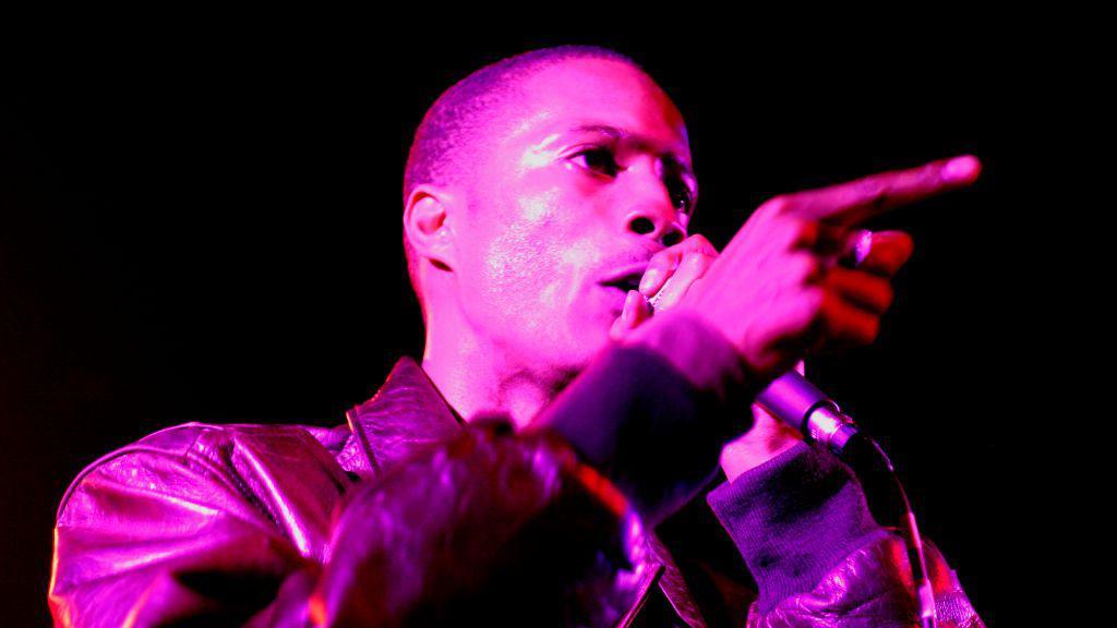 D Double E holds a microphone to his lips as he performs on stage. He is wearing a leather jacket and looking away to the right of the camera towards the audience. 