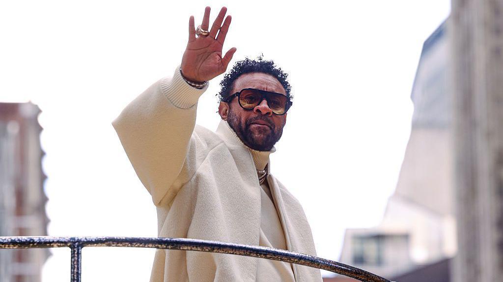 Shaggy wears a large, off-white woolen coat as he waves at a crowd below. He is standing on a raised platform or balcony, with tall buildings just visible in the background.