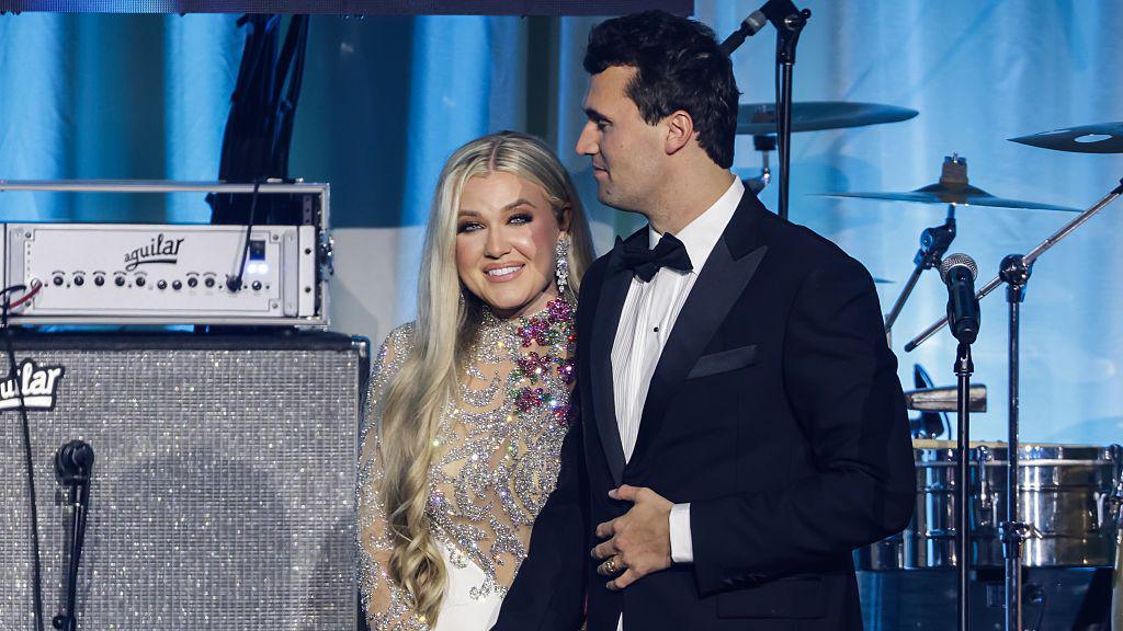 Charlie Kirk wears a black tuxedo in front of a bandstand and holds the hand of his wife, Erika Lane Frantzve, who is wearing a sparkling gown, during the Turning Point USA Inaugural-Eve Ball.