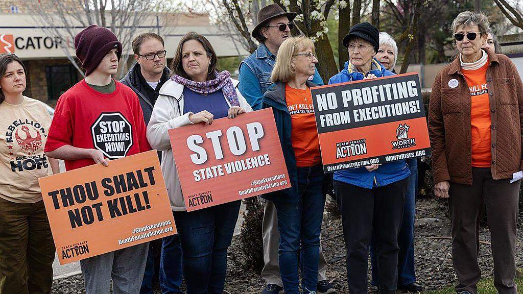 A group of protesters in coats and hats hold signs that say "stop state violence" and "thou shalt not kill".