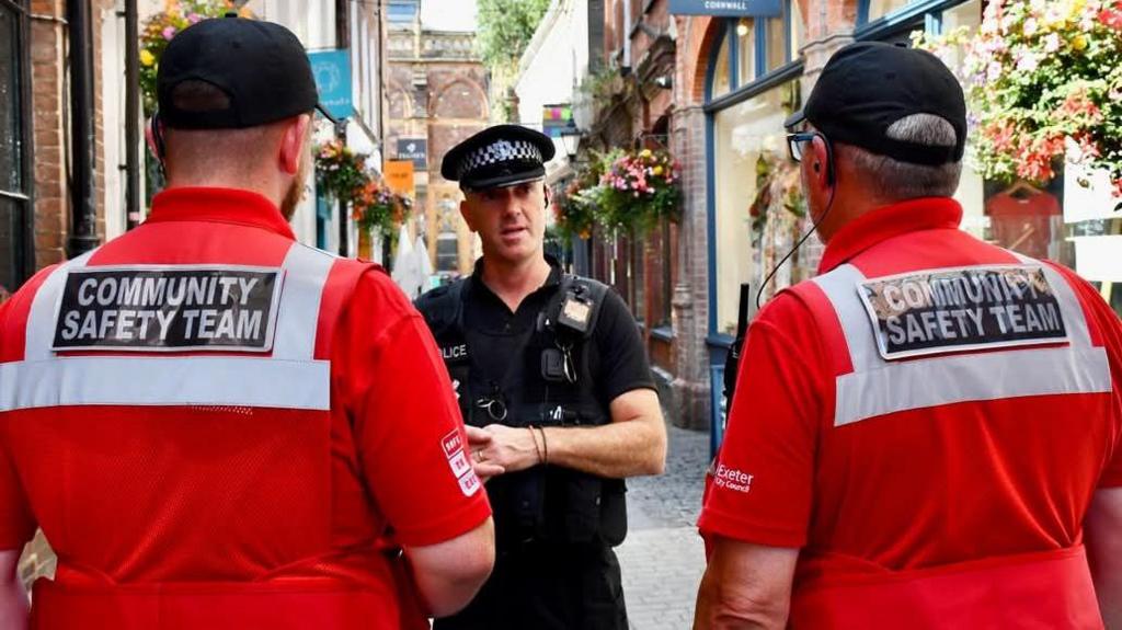 Two members of Exeter's Community Safety Team wearing red uniforms and black baseball caps look at a Devon and Cornwall Police officer wearing a black uniform while stood on Gandy Street in the city centre as they have a conversation. Several of the shops have hanging baskets filled with colourful flowers on the street.