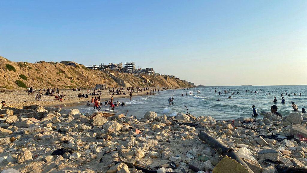 A beach and the sea, with concrete and rocks in the foreground. Lots of figures are in the sea, while damaged buildings can been seen on the headland above. The sky is blue.