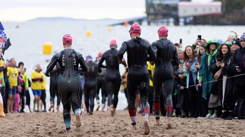 Two men and woman wearing wetsuits run towards the sea watched by spectators