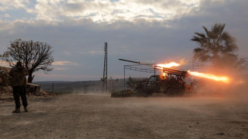 A rebel fighter covers his ears as a multi-barrel rocket launcher fires against government forces on the outskirts of the city of Hama, central Syria (4 December 2024)