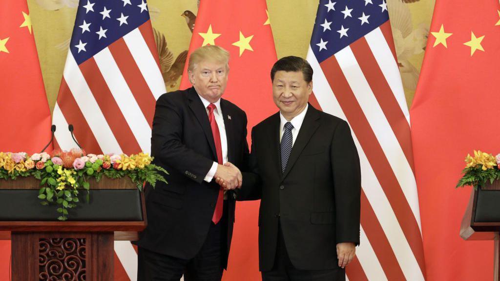 Donald Trump and Xi Jinping shake hands on a stage in front of large US and Chinese flags