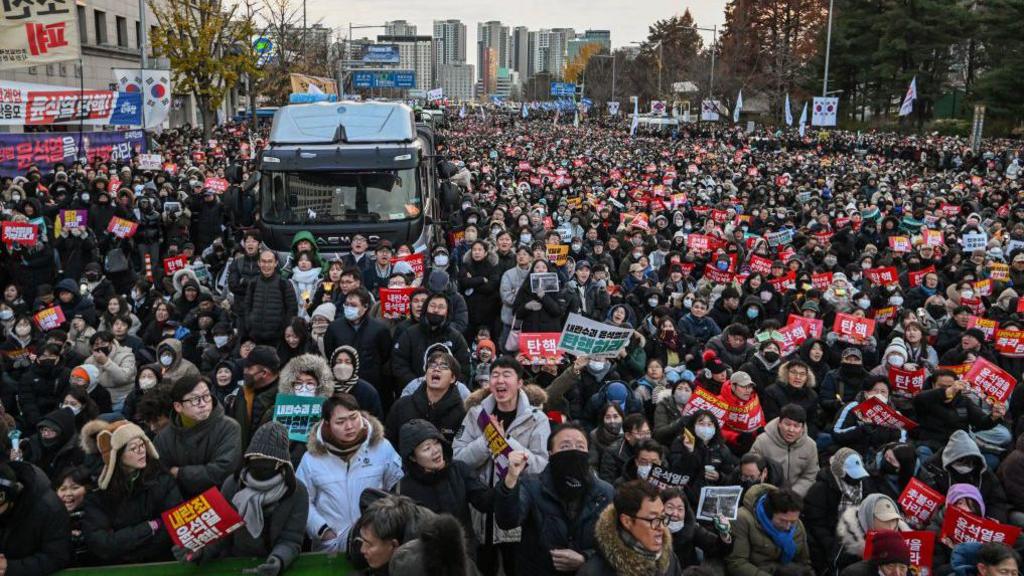 People take part in a protest calling for the ouster of South Korean President Yoon Suk Yeol outside the National Assembly in Seoul on December 7, 2024. Huge crowds in winter clothing stand with posters in hand and waving the Korean flag. In the middle is a dark van, likely a police vehicle.
