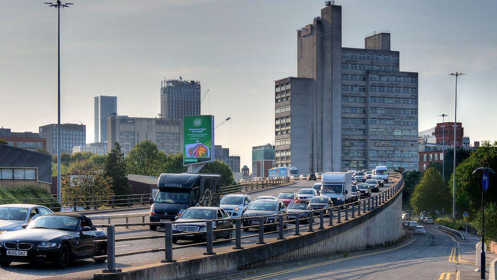 Cars line up along a section of the Mancunian way. The barriered road can be seen sloping upwards alongside a slip road, with city centre buildings seen rising up in the background. 