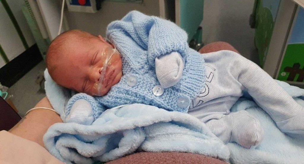 A baby boy dressed in baby blue clothing being held in a hospital.