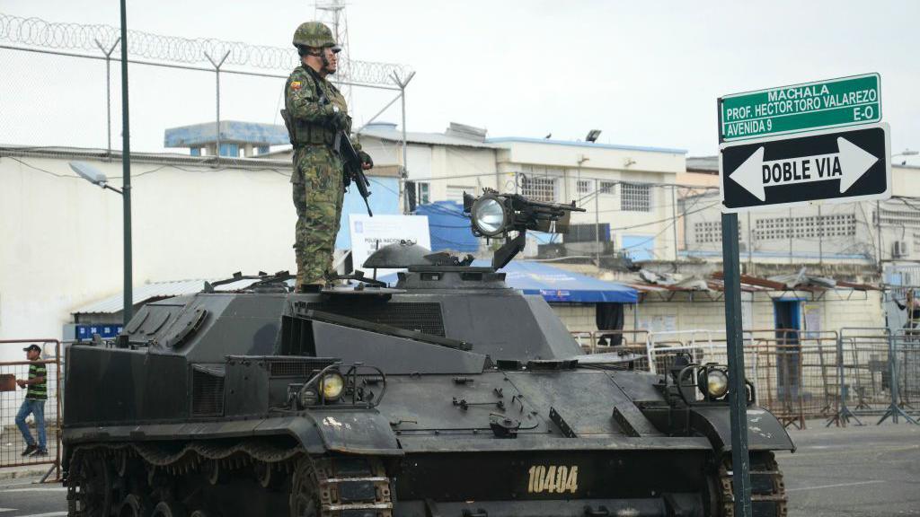Two Ecuadorean soldiers stand guard atop a tank outside Machala prison, in Machala, Ecuador on January 15, 2024. One is carrying a weapon, and in the background the wall of the prison can be seen with fencing and barbed wire on top.
