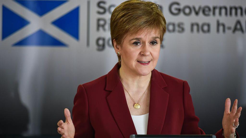 Nicola Sturgeon standing at a podium at a Covid briefing in March 2020. She is wearing a maroon jacket, gold necklace and a white top. There is a white background featuring a Saltire and the Scottish government logo.