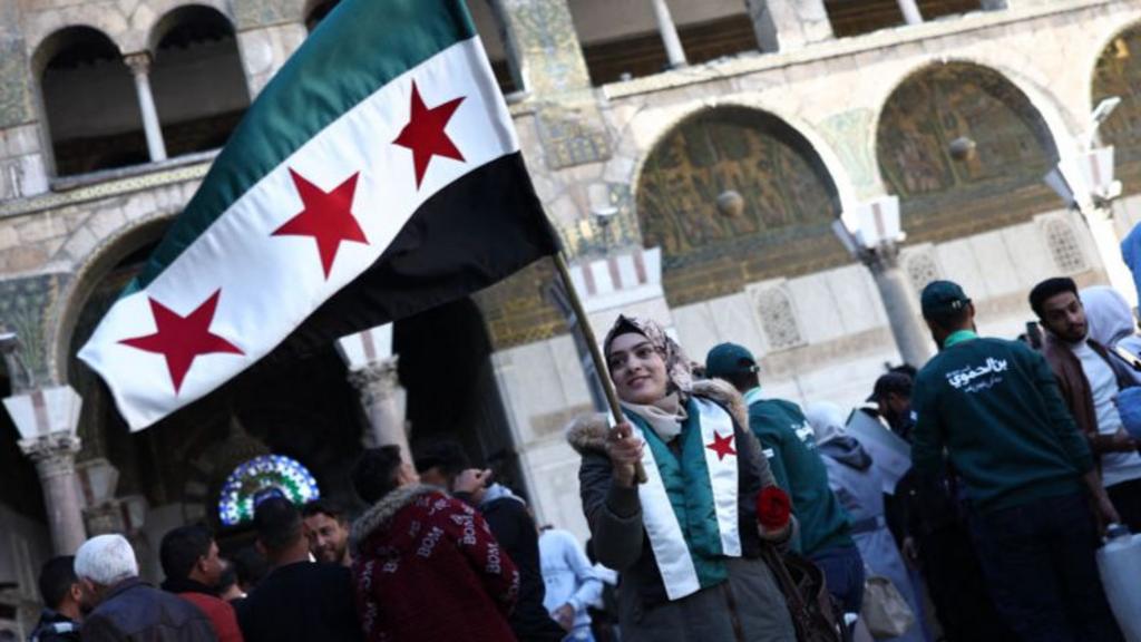 A Syrian woman waves the rebel's flag while wearing another as a scarf