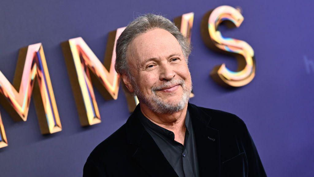 Actor Billy Crystal smiles as he is photographed a red carpet event. He has grey hair and a heard and wears a dark suit.