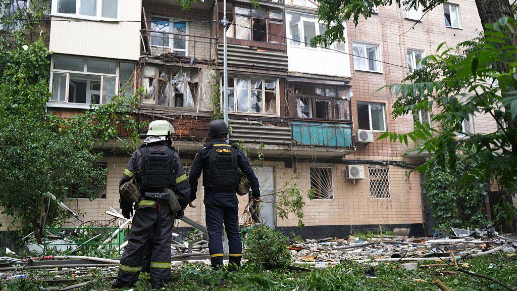 Two firefighters stand outside a damaged residential building after a drone and missile strike in Kharkiv, Ukraine. Both are wearing black uniforms and protective gear over their heads