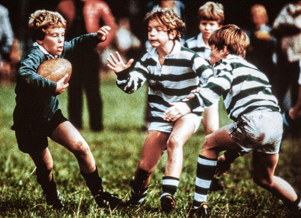 Three boys playing rugby. One is holding the ball in dark green while two opponents in green and white hoops try and tackle him.