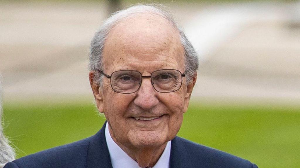 George Mitchell is smiling at the camera. He has short, balding grey hair and is wearing black-rimmed glasses. He is an older man with a lined face. He is wearing a blue suit jacket and white shirt. In the background, out of focus, we can see a lawn and possibly a road. It is daytime.