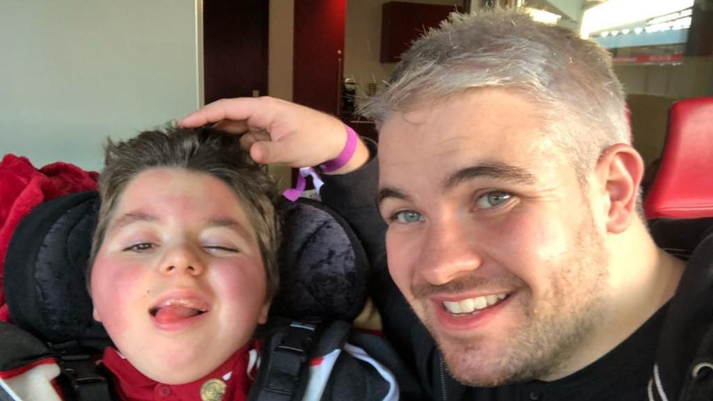 Paul Woodward is sitting next to his son Kalel with his hand resting on his head. Kalel is sticking his tongue out while Paul is smiling. 