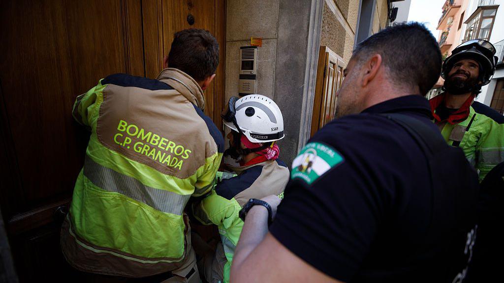 Three firefighters in fluorescent jackets attempt to enter a building through a wooden door while a police officer dressed in black watches on.