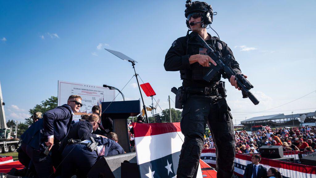 A police officer with a helmet, sunglasses and a large gun standing above a crowd of people crouching behind a podium