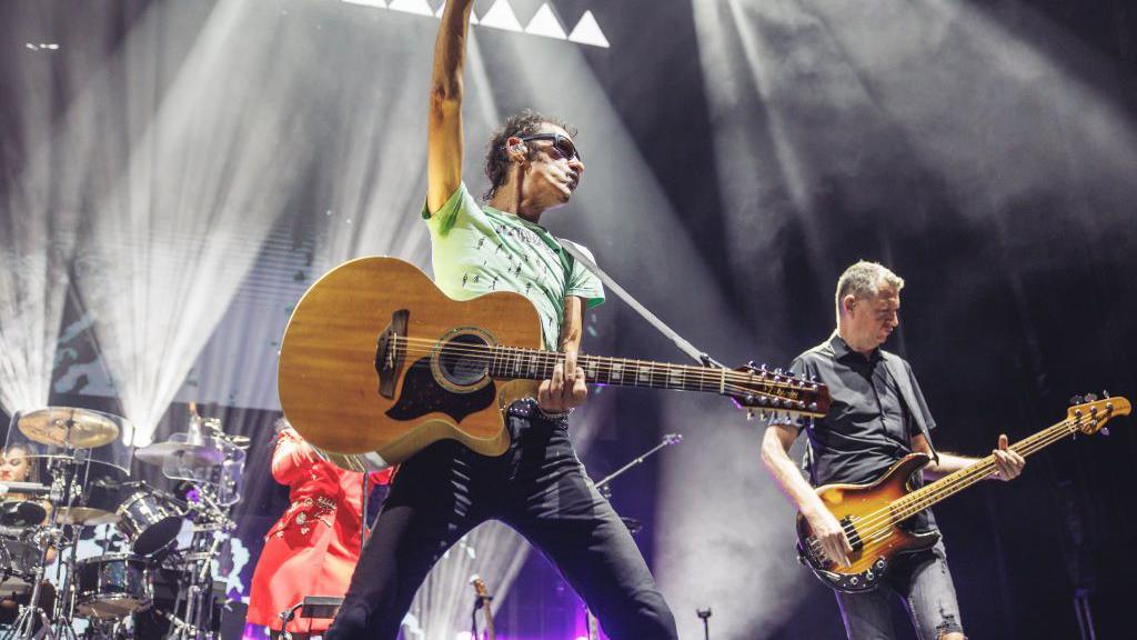 A guitarist wearing a green t-shirt and black skinny jeans with his arm up in the air, holding a guitar. A man wearing a black shirt and jeans is in the background holding a guitar. They are both stood on a stage. Spotlights are shining and there are drums in the background.