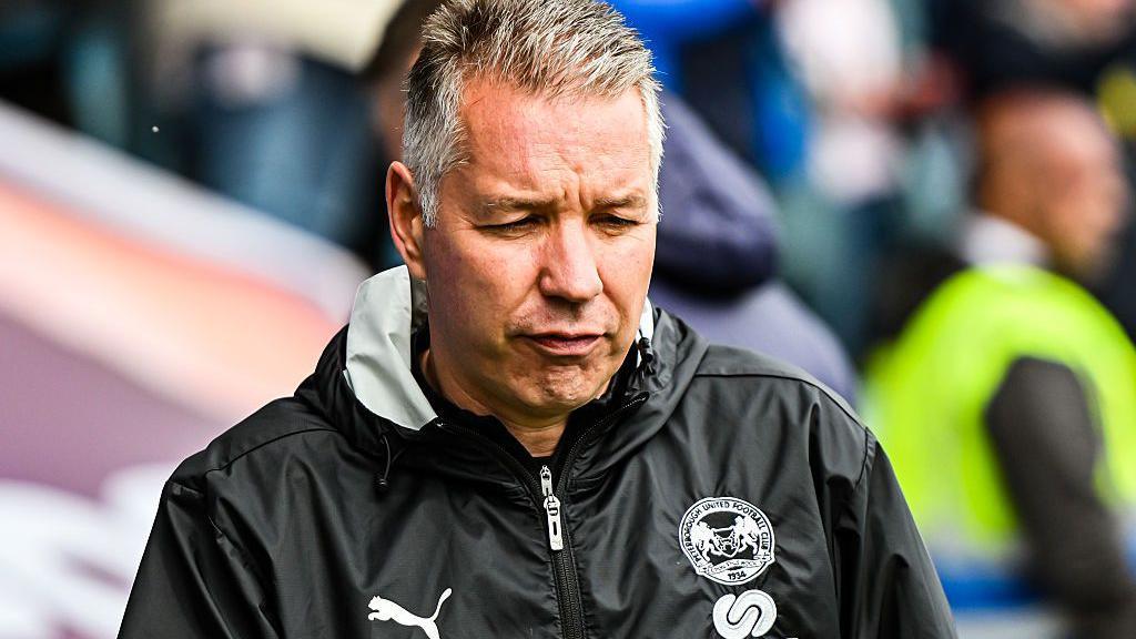 Darren Ferguson frowning and looking downwards. He is wearing a black rain jacket with a Puma logo and others.