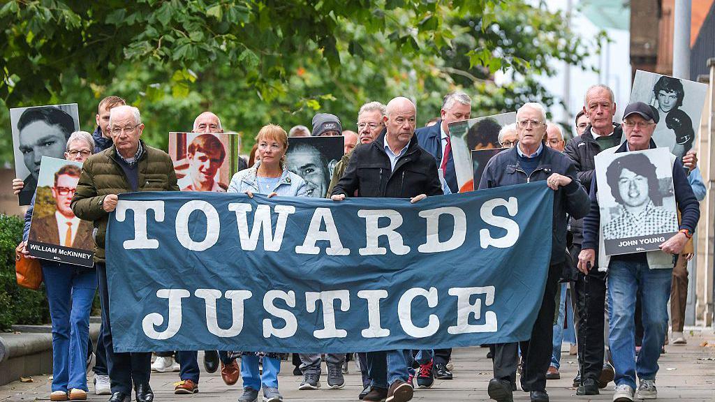 Families and supporters of those who were killed walk in a group holding a blue banner with white writing saying 'Towards Justice'. They are also holding black and white, and coloured images of the victims. There are trees above their heads as they walk along the street.