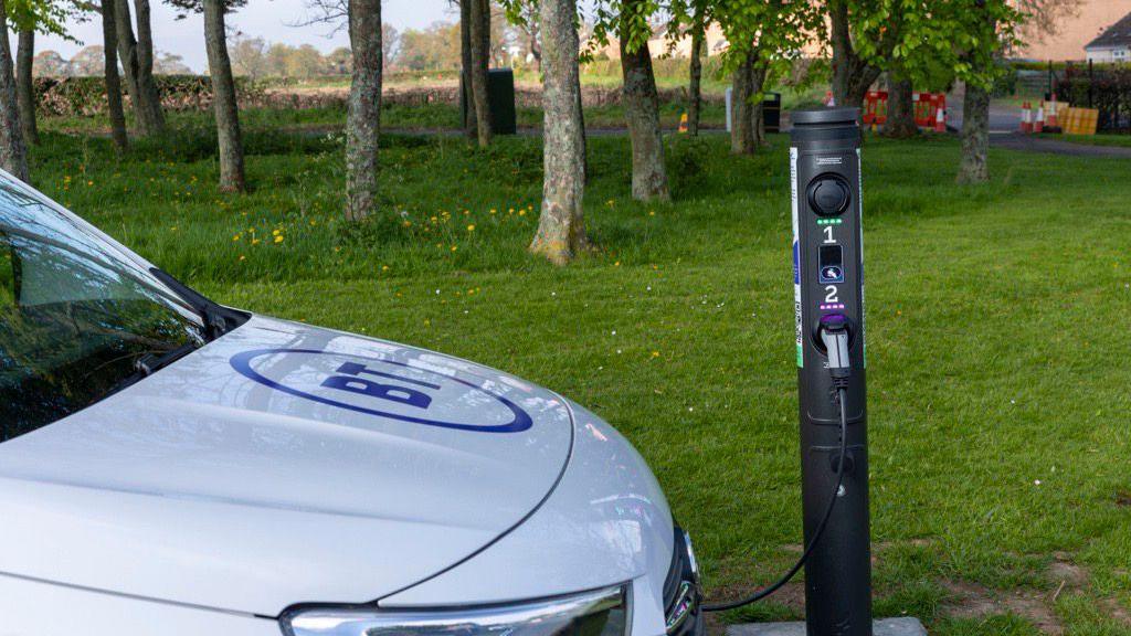 A white car with the BT logo on its bonnet, parked next to a black tower-shaped charging point, with trees and grass in the background.