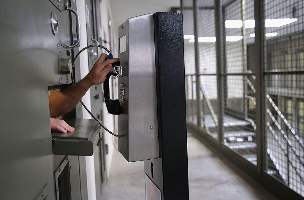 An arm reaches out from a cell inside the Adelanto immigrant detention centre to reach for a pay phone. A stairway in the background is covered in a cage and the cell only has a small opening for the arm to reach out.