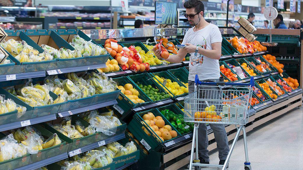 A man wearing a t-shirt uses a hand-held device to scan a net of oranges as he shops in the fruit and vegetable aisle of a Tesco supermarket