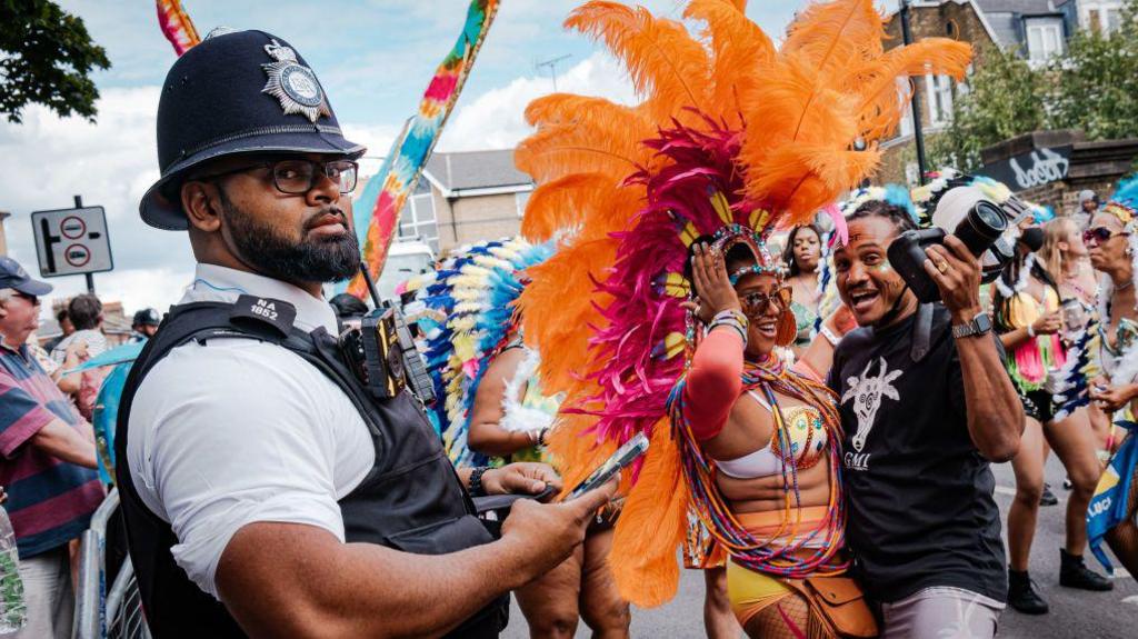 A police officer in uniform stands watch at Notting Hill Carnival, while a woman in a colourful feathered costume poses for a photo with a man holding a camera. Other costumed performers and onlookers can be seen in the background.