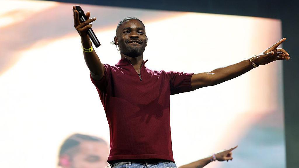 A man in a purple t-shirt with a collar and zip-up neck holds a microphone as he points with both hands at the crowd out in front of him. In the background a large, bright screen shows a live feed of the on-stage performance.
