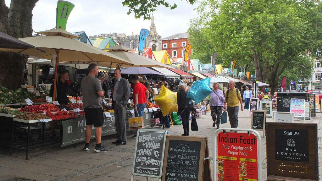 Shoppers on Norwich Market. A boards advertising stalls are in the foreground