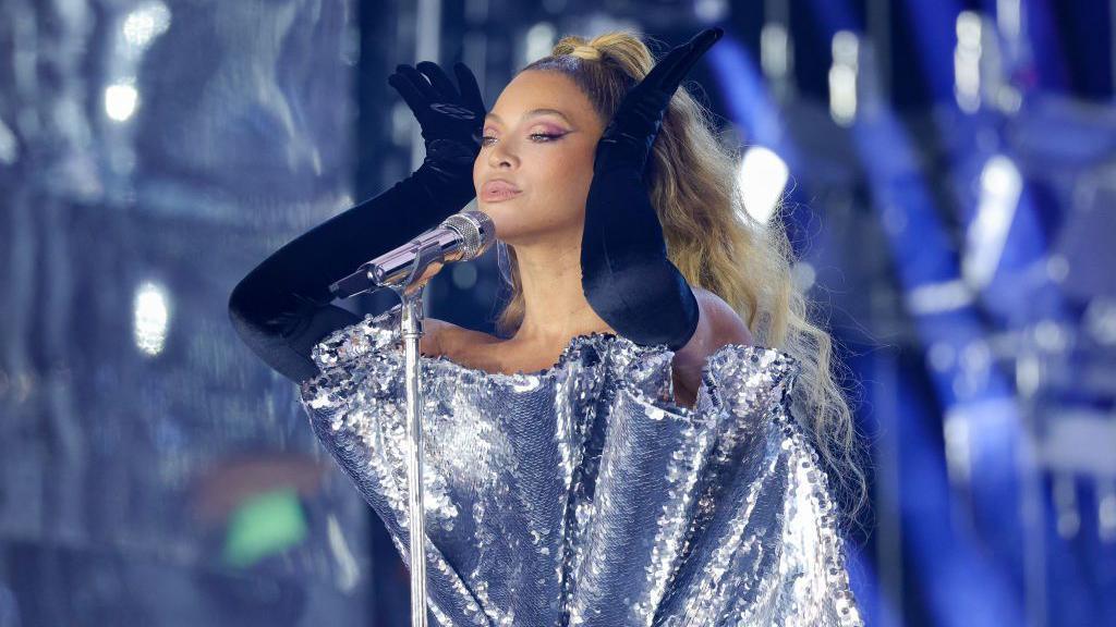 Beyoncé holds her hands to her head, while wearing a ruffled, metallic dress on stage at her Renaissance World Tour