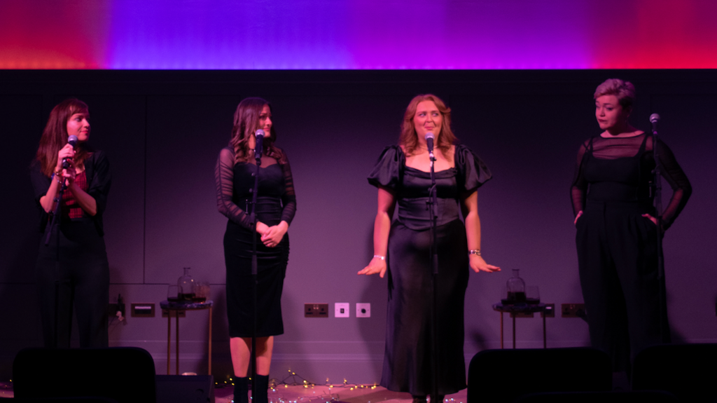 Four women stand together on a stage, in front of microphones. They are all dressed elegantly in black outfits. 