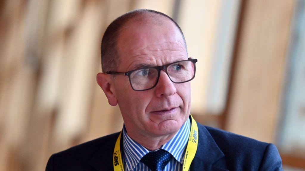 a balding man with short cropped hair is wearing glasses, a blue jacket, blue and white striped shirt and blu tie