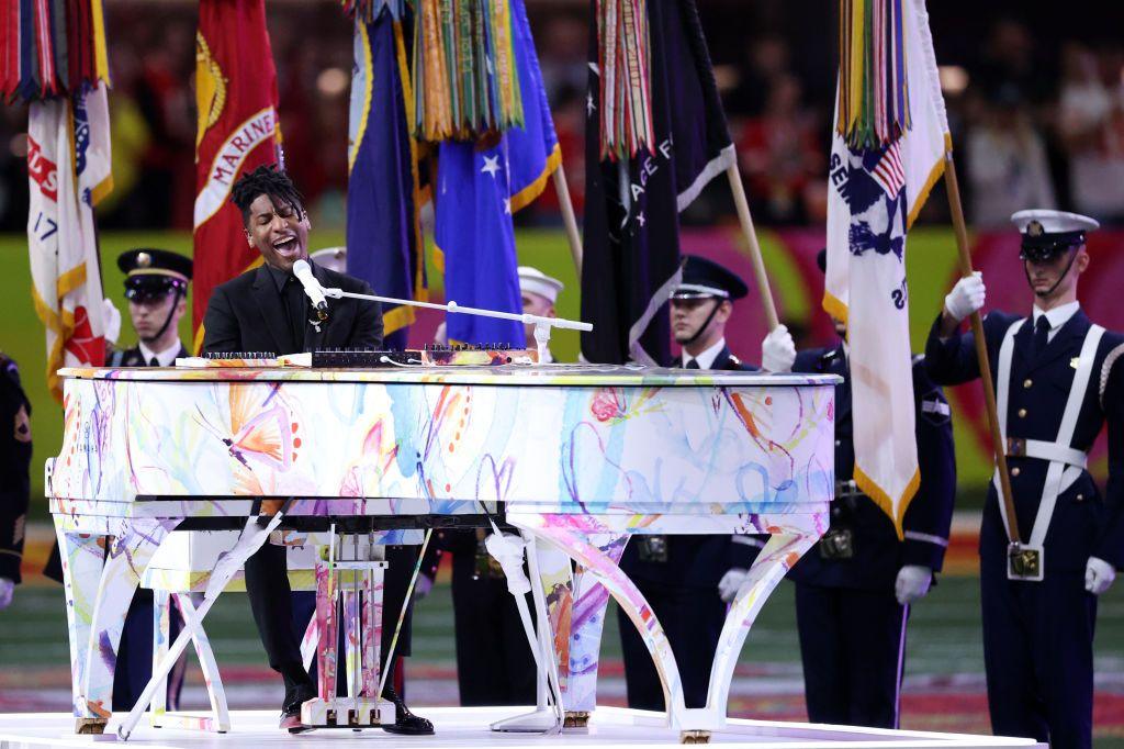 Jon Batiste sits at a technicolor grand piano, flanked by members of the US Air Force, who are holding flags