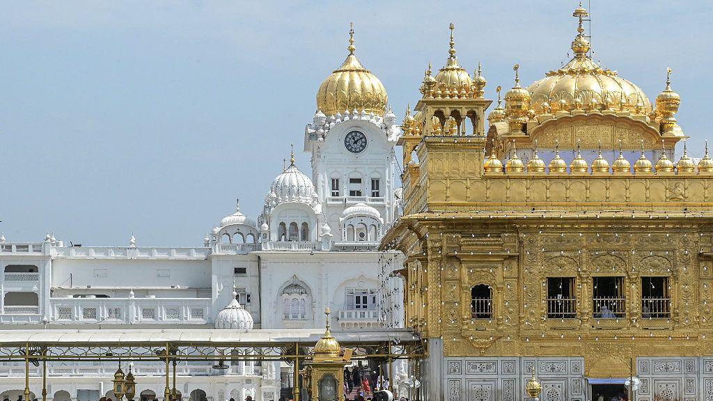 A view of the Golden Temple complex in Amritsar, Punjab. The gold-covered Harmandir Sahib dominates the foreground, while white marble buildings with domes and a clock tower rise behind it under a clear blue sky. The temple is the holiest shrine in Sikhism and is visited by millions of pilgrims each year.