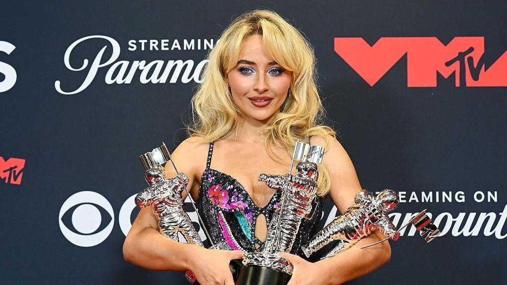 Sabrina Carpenter holding three awards.