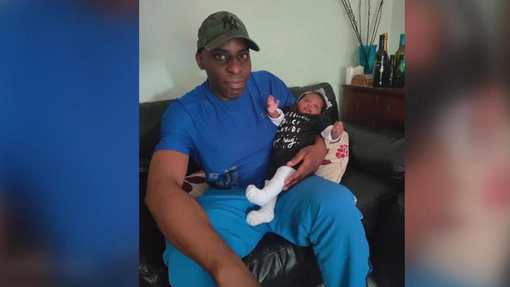 A man sitting on a settee with a small baby. He is wearing a green baseball cap, a blue t-shirt and blue trousers. 