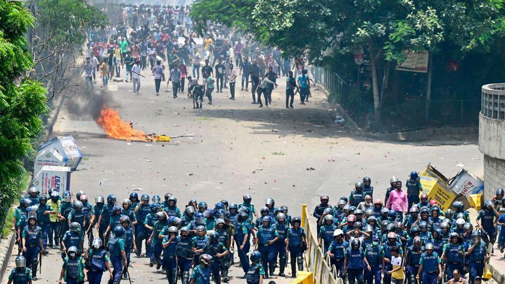 A group of police in blue uniforms and helmets, with their backs facing a crowd of protesters. A burning object lies on the ground between them.