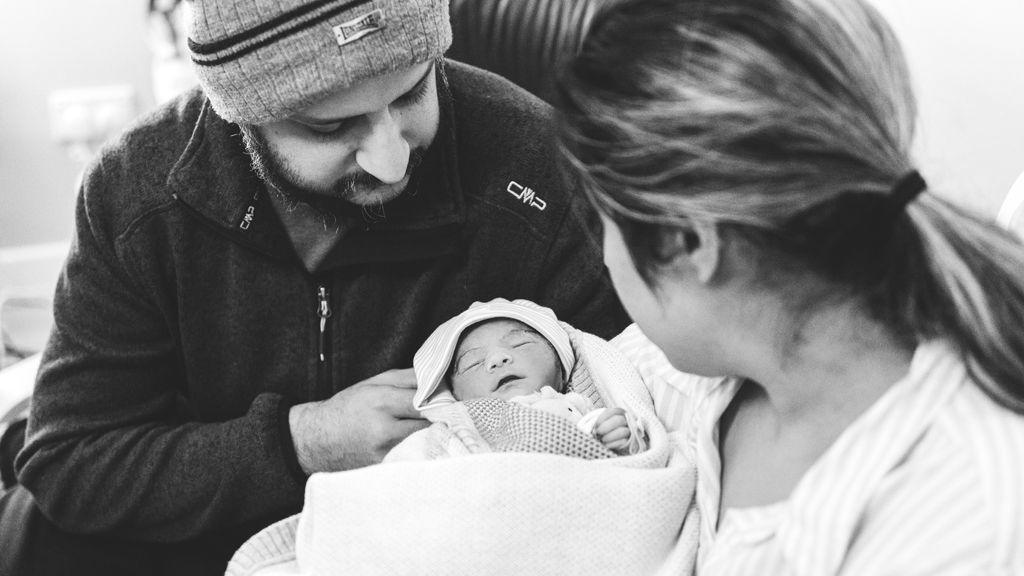 Black and white photo of Amarjit and Mandip holding their newborn baby Asees. She is wrapped in a blanket with a hat on. She is in Amarjit's arms and both parents are looking down at her. 