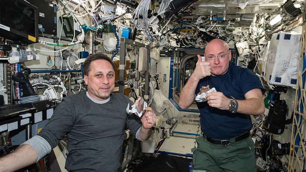 Two male astronauts posing for picture in the ISS, floating and eating snacks. There is lots of machinery around them.